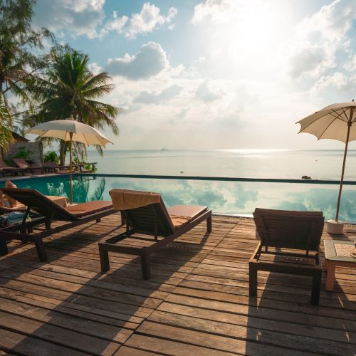 A serene beach scene with lounge chairs, umbrellas, and palm trees overlooking a calm sea with a bright sky and scattered clouds.