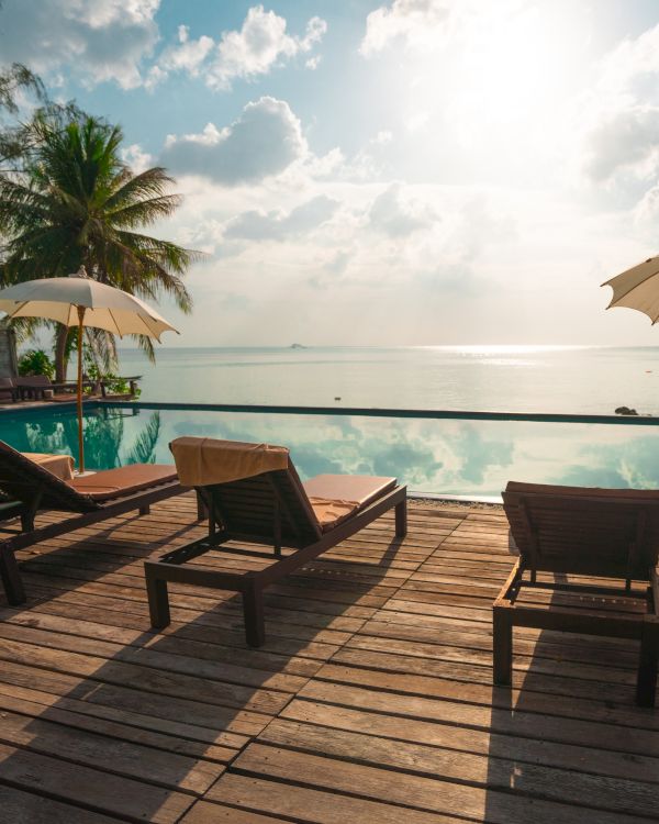 A serene beach scene with lounge chairs, umbrellas, and palm trees overlooking a calm sea with a bright sky and scattered clouds.