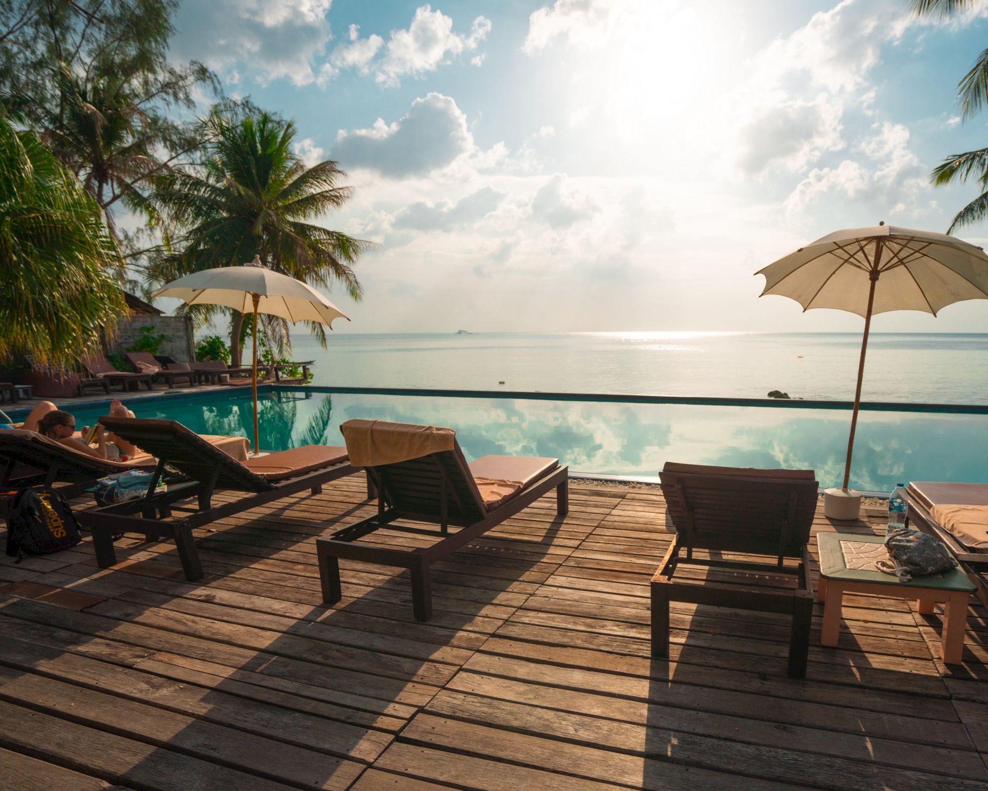 Lounge chairs and umbrellas by a pool overlook a calm sea, with palm trees and a bright sky adding to the tranquil scenery.