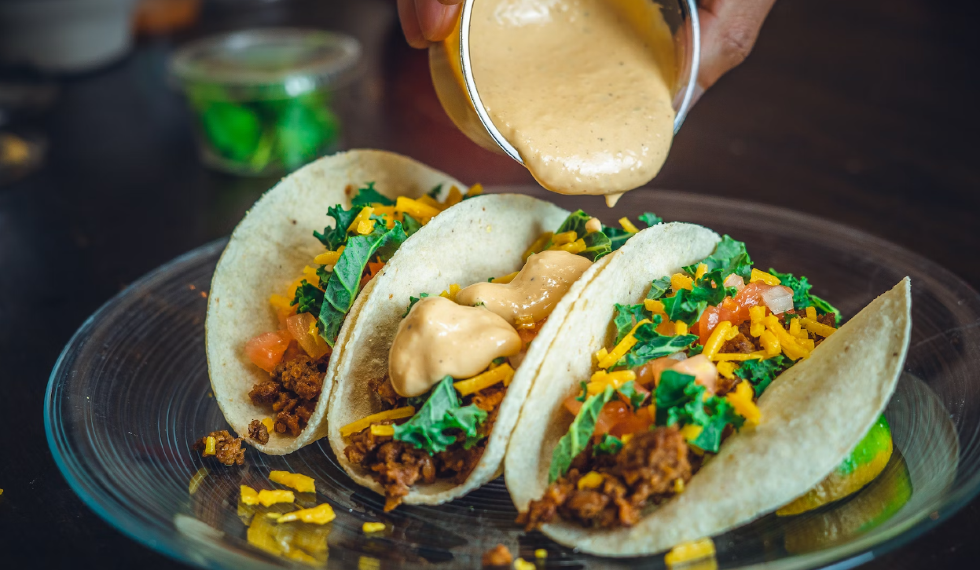 Three tacos with toppings like lettuce, cheese, and sauce being poured, on a plate.