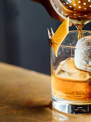 A cocktail is being poured into a glass with ice and a lemon twist garnish on a countertop.
