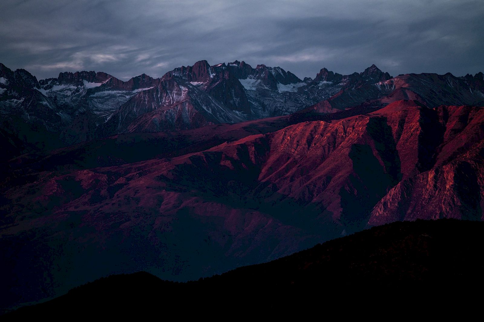 A mountain range bathed in dramatic red light under a cloudy sky.