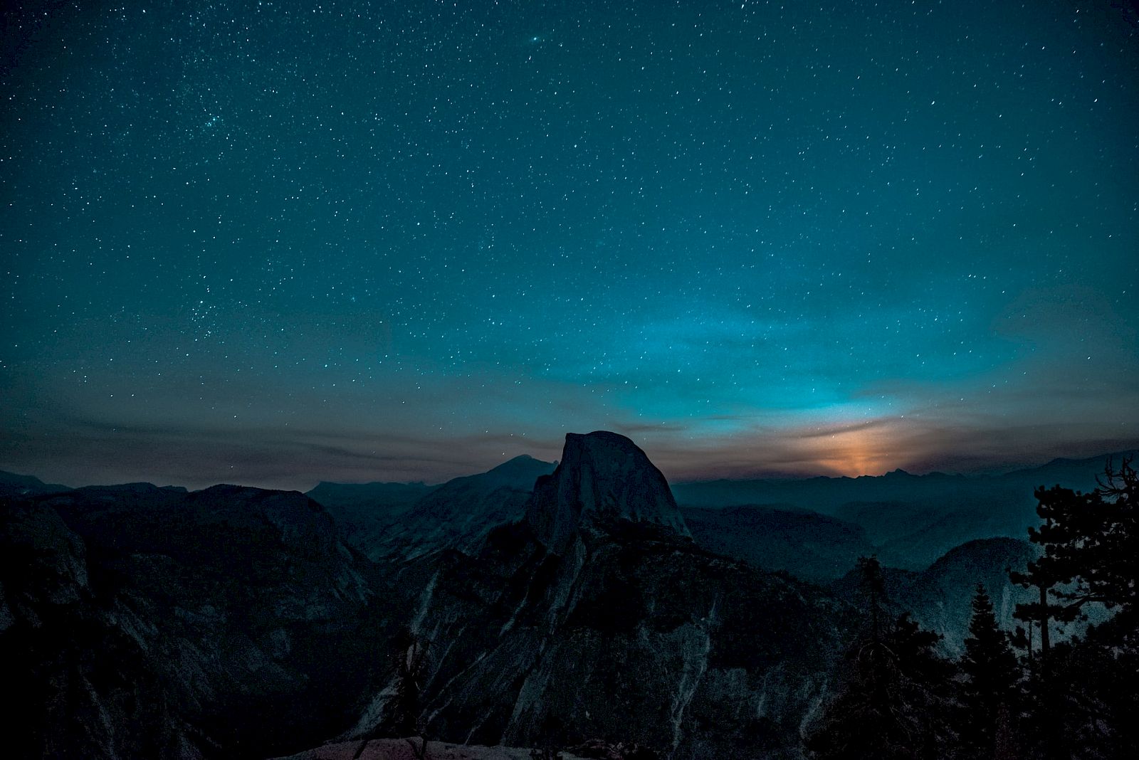 A starry night sky over a mountain landscape with a glowing horizon, trees silhouetted in the foreground.