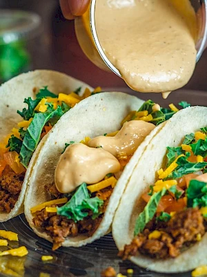 Three tacos filled with ground meat, cheese, lettuce, and tomatoes are being topped with a creamy sauce on a plate.
