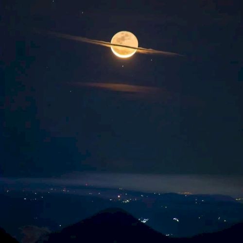 The image shows a full moon with thin clouds creating a Saturn-like ring illusion, set against a dark sky above a faintly lit landscape.