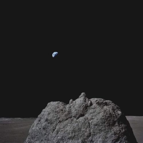 The image shows the Earth rising above the lunar horizon, with a rocky surface in the foreground against a dark sky.