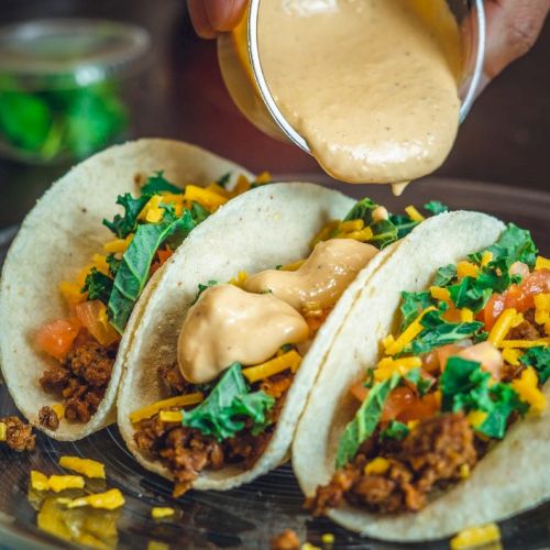 Three tacos with ground meat, cheese, lettuce, and tomatoes, being topped with a creamy sauce on a glass plate.