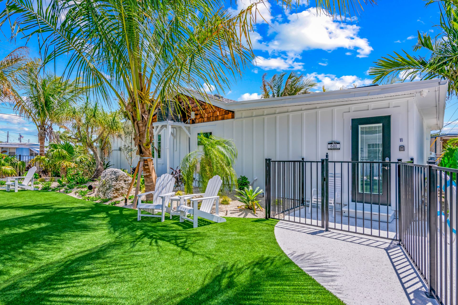 A small white house with a fenced entrance, surrounded by green lawn and palm trees, under a bright blue sky with fluffy clouds.