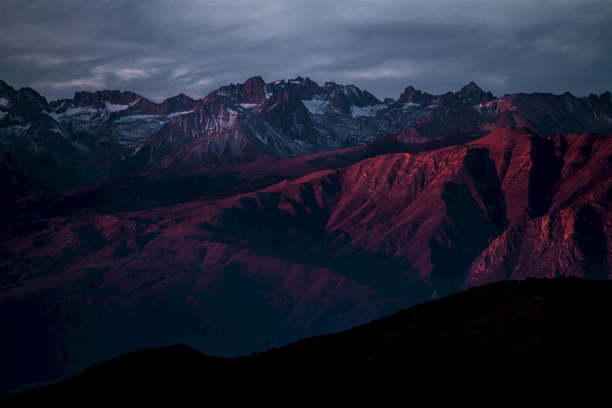 A range of mountains with a dramatic red tint from the setting sun under a cloudy sky.