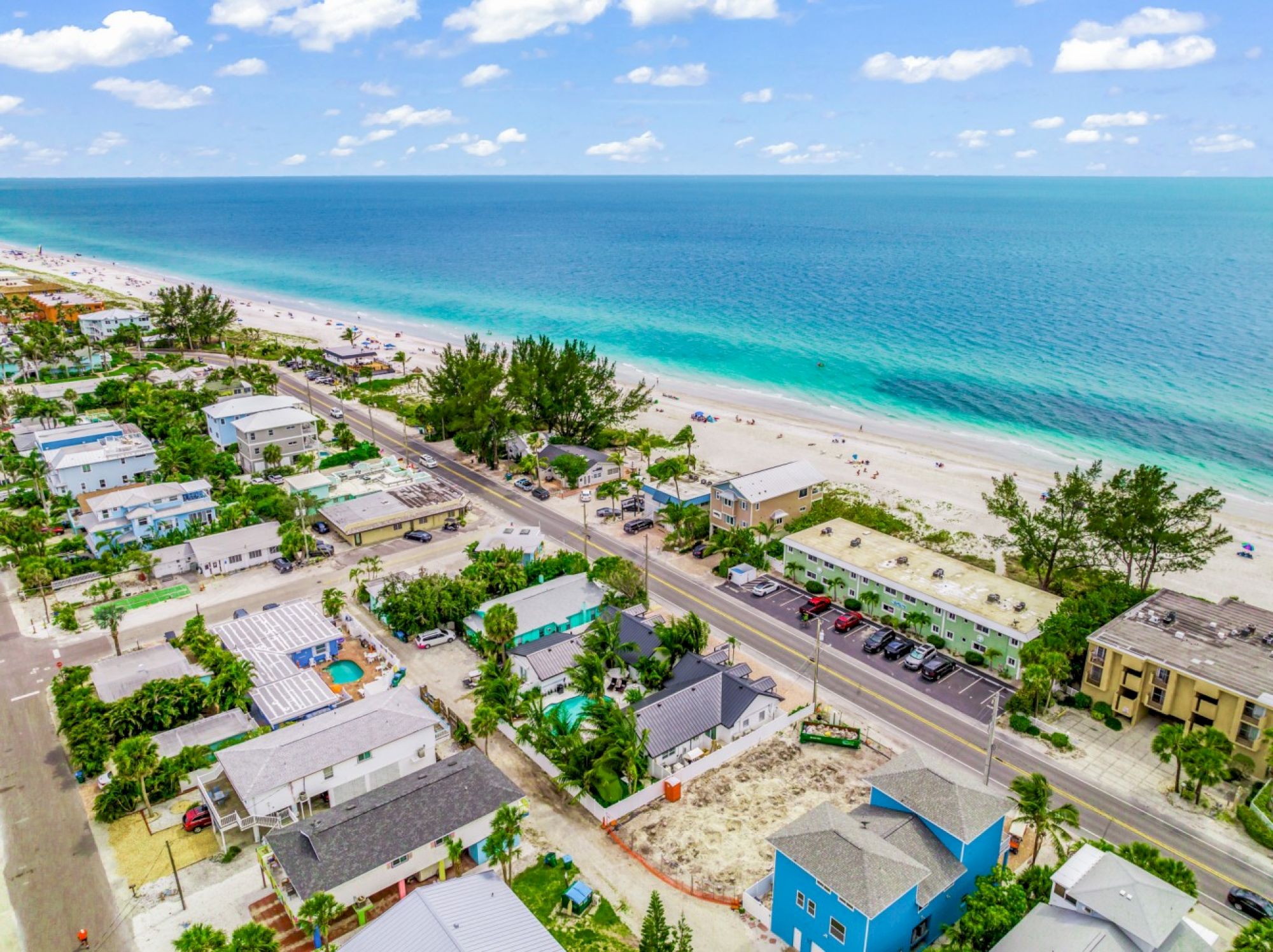An aerial view of a coastal area with beachfront properties, a road, parked cars, and a clear ocean under a blue sky with clouds.