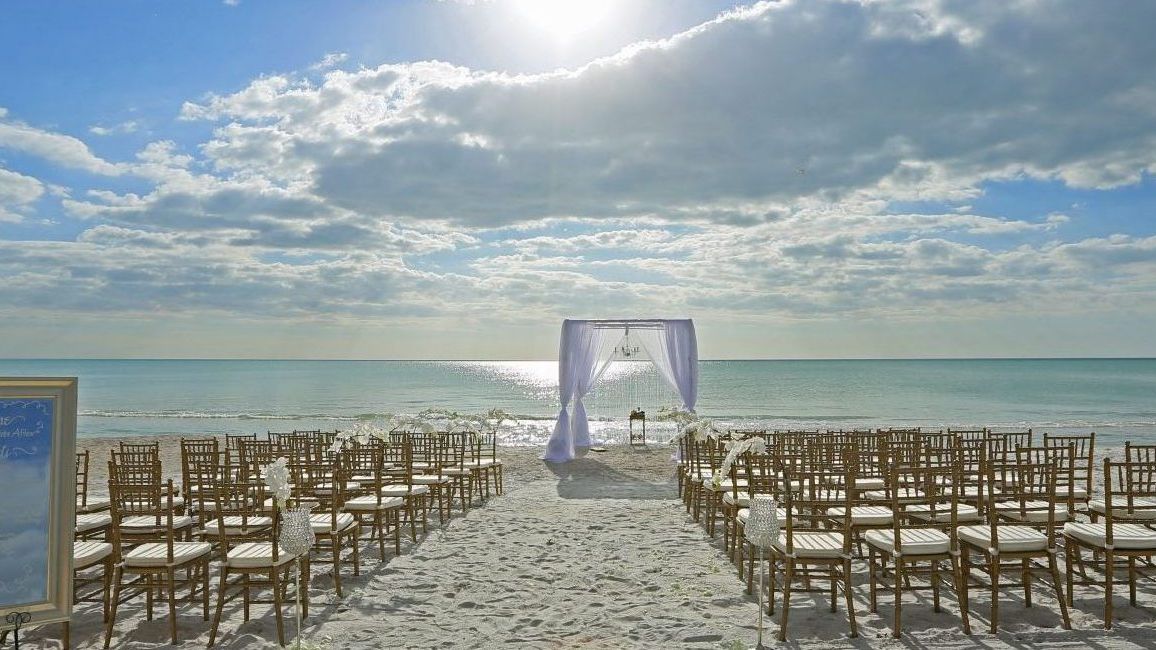 A beach wedding setup with an arch, chairs, and a welcome sign, under a blue sky with clouds and sunlight.