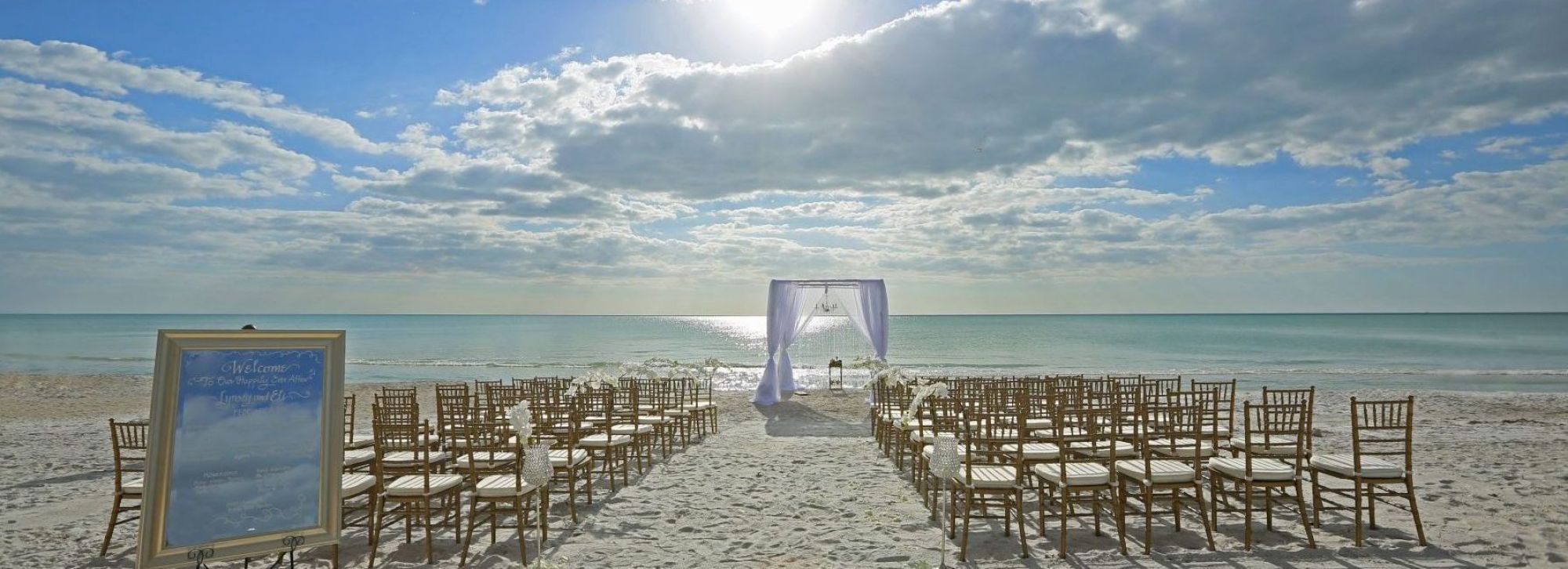 A beach wedding setup with an arch, chairs, and a welcome sign, under a blue sky with clouds and sunlight.