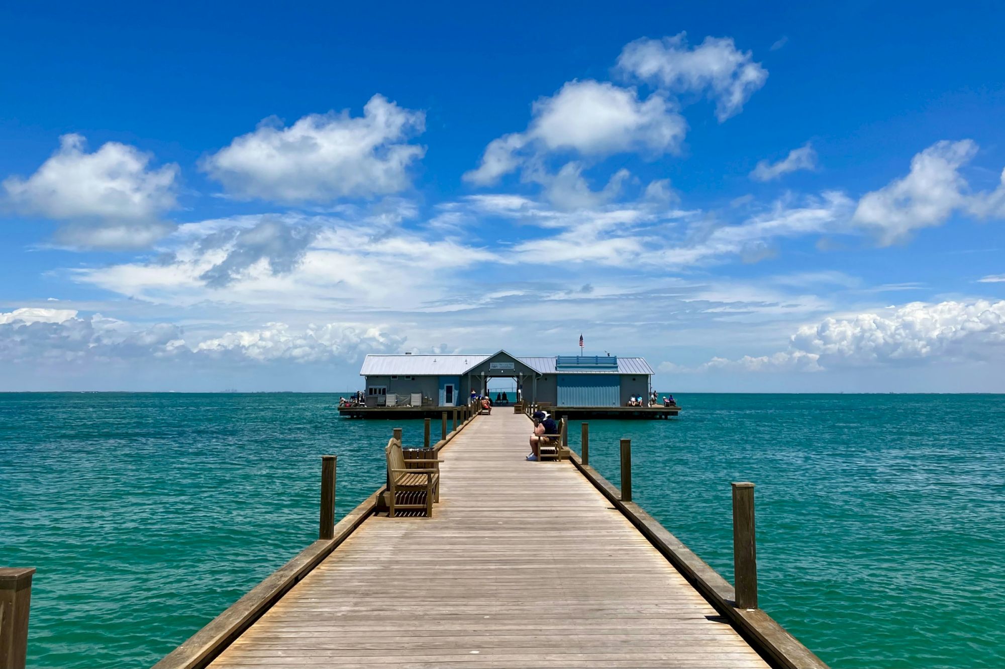 A wooden pier extends over a calm ocean, leading to a building under a bright blue sky with fluffy clouds.