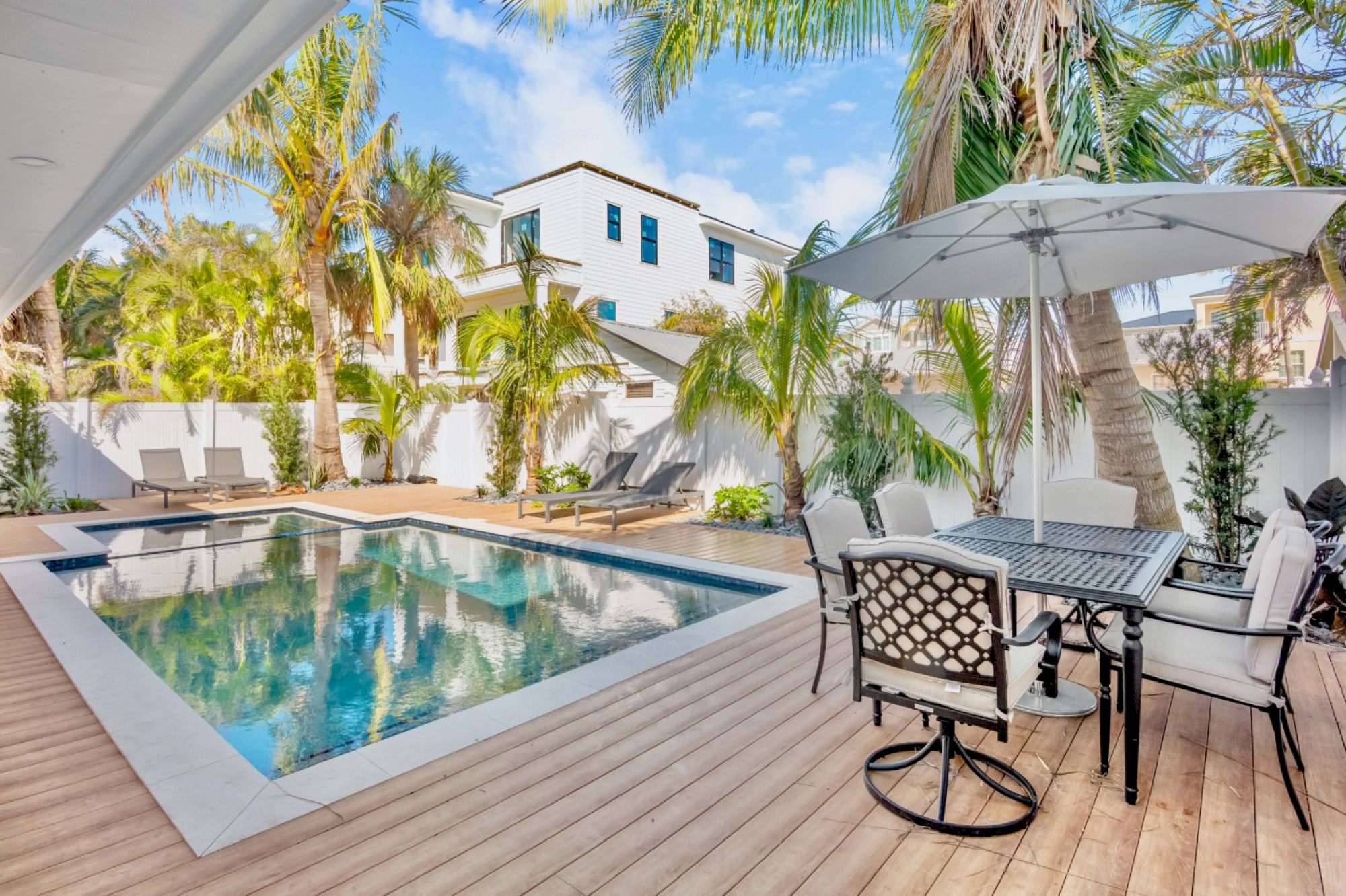 An outdoor area with a pool, surrounded by palm trees, deck chairs, and a dining set under an umbrella on a wooden deck.