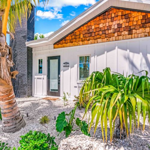 The image shows a house with a modern facade, surrounded by tropical plants and a palm tree, under a bright blue sky.