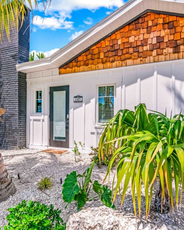The image shows a house with a modern facade, surrounded by tropical plants and a palm tree, under a bright blue sky.