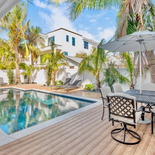 A stylish outdoor area featuring a pool, lounge chairs, palm trees, and a dining set under an umbrella on a wooden deck.
