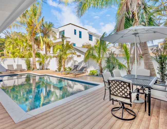A stylish outdoor area featuring a pool, lounge chairs, palm trees, and a dining set under an umbrella on a wooden deck.