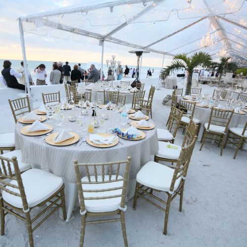 The image shows an elegant beachside event setup with tables and chairs under a clear canopy, overlooking the ocean at sunset.