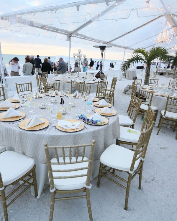 The image shows an elegant beachside event setup with tables and chairs under a clear canopy, overlooking the ocean at sunset.