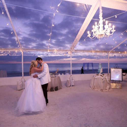 A couple is dancing under a decorated canopy with string lights, overlooking the ocean at sunset.