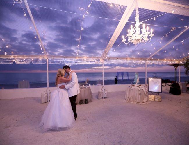 A couple is dancing under a decorated canopy with string lights, overlooking the ocean at sunset.