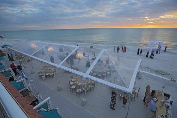 A beachside event with guests under a clear tent, tables, and a sunset backdrop. People are also gathered near the shore.