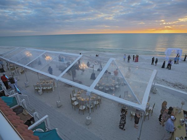 A beachside event with guests under a clear tent, tables, and a sunset backdrop. People are also gathered near the shore.
