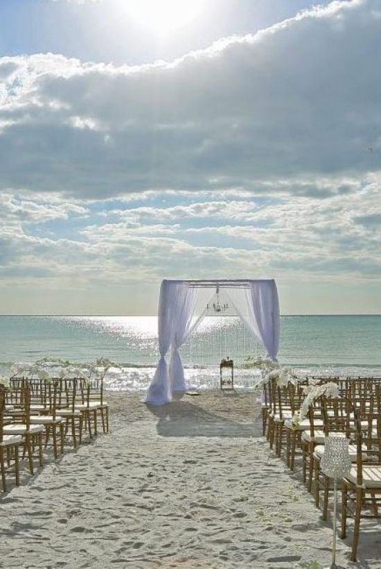 A beach wedding setup with an archway, multiple rows of chairs facing the ocean, and a framed sign on the sand under a cloudy blue sky.