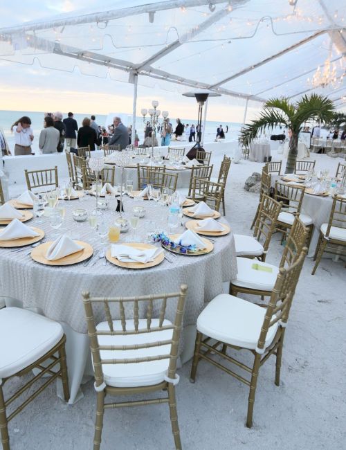 The image shows a beachside event setup with round tables, chairs, and a clear tent, offering a scenic view of the ocean.