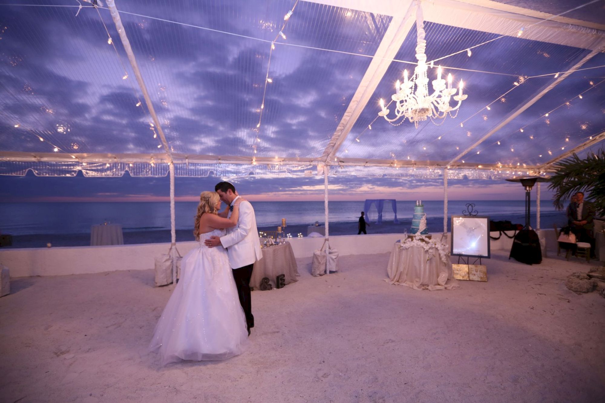 A couple dances under a decorated tent by the sea at dusk. String lights and a chandelier illuminate the sandy beach setting.