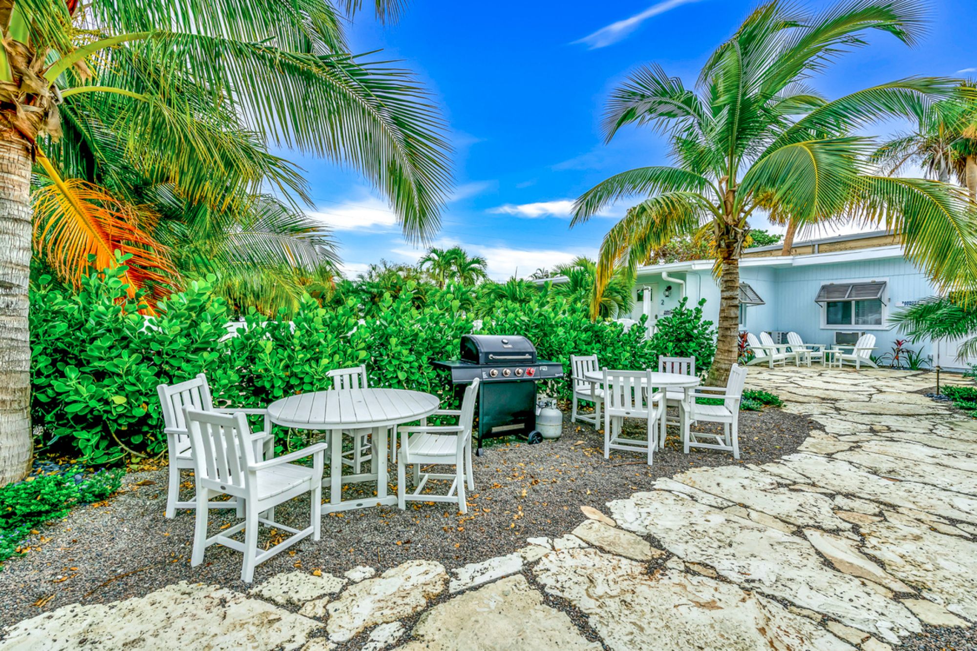 A backyard patio with white chairs, tables, a grill, palm trees, and lush greenery under a partly cloudy blue sky.