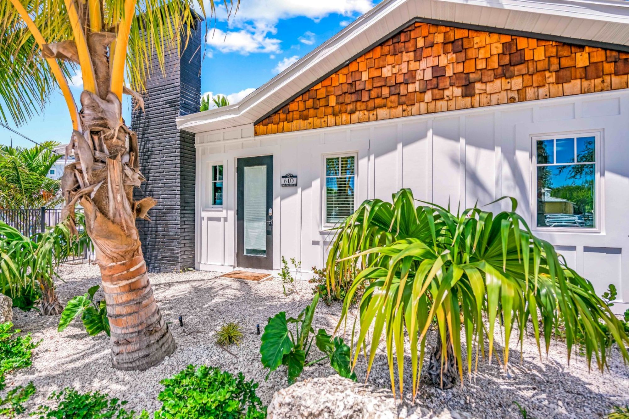 A charming house with wooden shingles, white walls, and lush tropical vegetation, set against a bright blue sky.