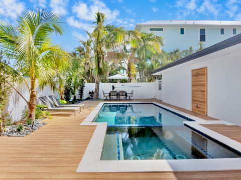 A backyard with a swimming pool, wooden deck, lounge chairs, palm trees, and a seating area under a clear blue sky.