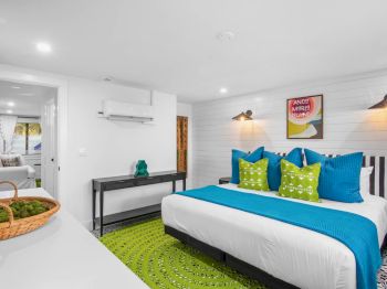 A bright bedroom with a large bed, blue and green accent pillows, a green rug, and artwork on the wall, creating a fresh, airy ambiance.