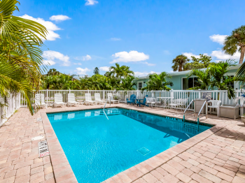 A clear swimming pool surrounded by a white fence, palm trees, and patio furniture, under a blue sky with scattered clouds.