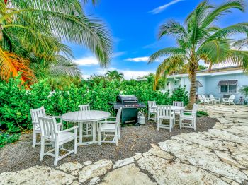 A patio with white chairs and tables, a barbecue grill, surrounded by lush greenery and palm trees under a bright blue sky.
