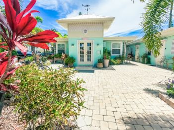 A pastel-colored house with a paved courtyard, surrounded by tropical plants, under a bright blue sky.