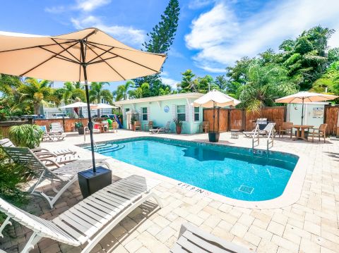 A serene pool area with lounge chairs, umbrellas, and a small building, surrounded by lush greenery and a clear blue sky.