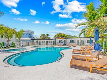 A serene pool area with wooden loungers, umbrellas, palm trees, and a clear blue sky, perfect for relaxation and enjoyment.