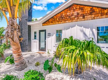 A small house with a shingle accent, surrounded by tropical plants and palm trees, under a bright blue sky and fluffy clouds.