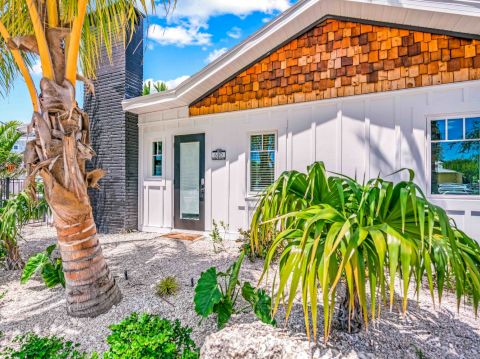 A small house with a shingle accent, surrounded by tropical plants and palm trees, under a bright blue sky and fluffy clouds.