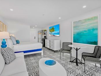 A modern bedroom with a bed, sofa, chairs, table, and vibrant artwork on the walls, decorated in white and blue accents.