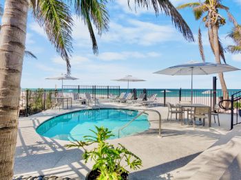 A poolside area with palm trees, lounge chairs, and umbrellas, adjacent to a beach with ocean views under a blue sky.