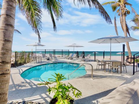 A poolside area with palm trees, lounge chairs, and umbrellas, adjacent to a beach with ocean views under a blue sky.