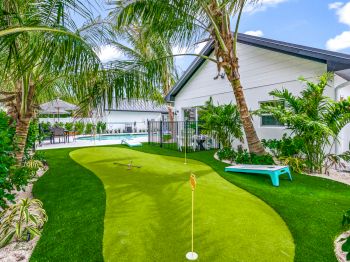 A backyard with a mini putting green, surrounded by palm trees, leads to a house and pool area, under a clear blue sky.