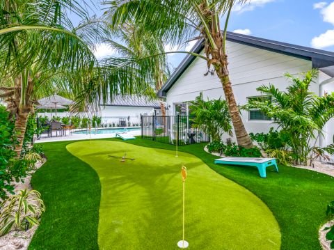 A backyard with a mini putting green, surrounded by palm trees, leads to a house and pool area, under a clear blue sky.