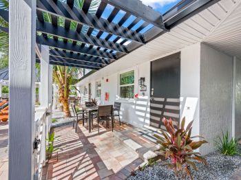 The image shows a patio with a pergola, outdoor furniture, and various plants next to a white building, creating a relaxing atmosphere.