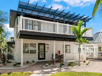 A two-story house with a pergola, white balcony, and tropical landscaping, alongside a gazebo and palm trees.
