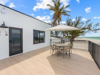 A patio with a table, chairs, and umbrella overlooks the beach. There are palm trees and a clear blue sky in the background.